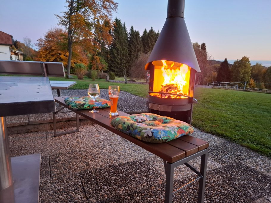 Außenkaminofen Gemütliche Terrasse mit Feuerstelle, Getränken und Kissen bei Dämmerung.